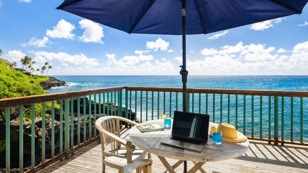 A laptop, book, water glass, and hat sit on a round table under a large blue umbrella on a wooden deck overlooking the ocean with a clear sky.